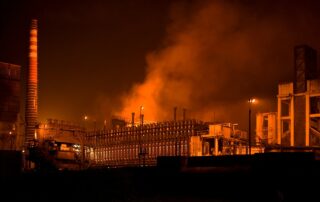 Eine Fabrik in der Nacht. Das Licht wirkt rötlich und Abgase sind in der Luft zu sehen.